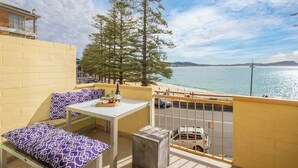 A balcony with a table and seating, overlooking a body of water and a road. - Oceans at Terrigal, a Beachfront Oasis (Terrigal)