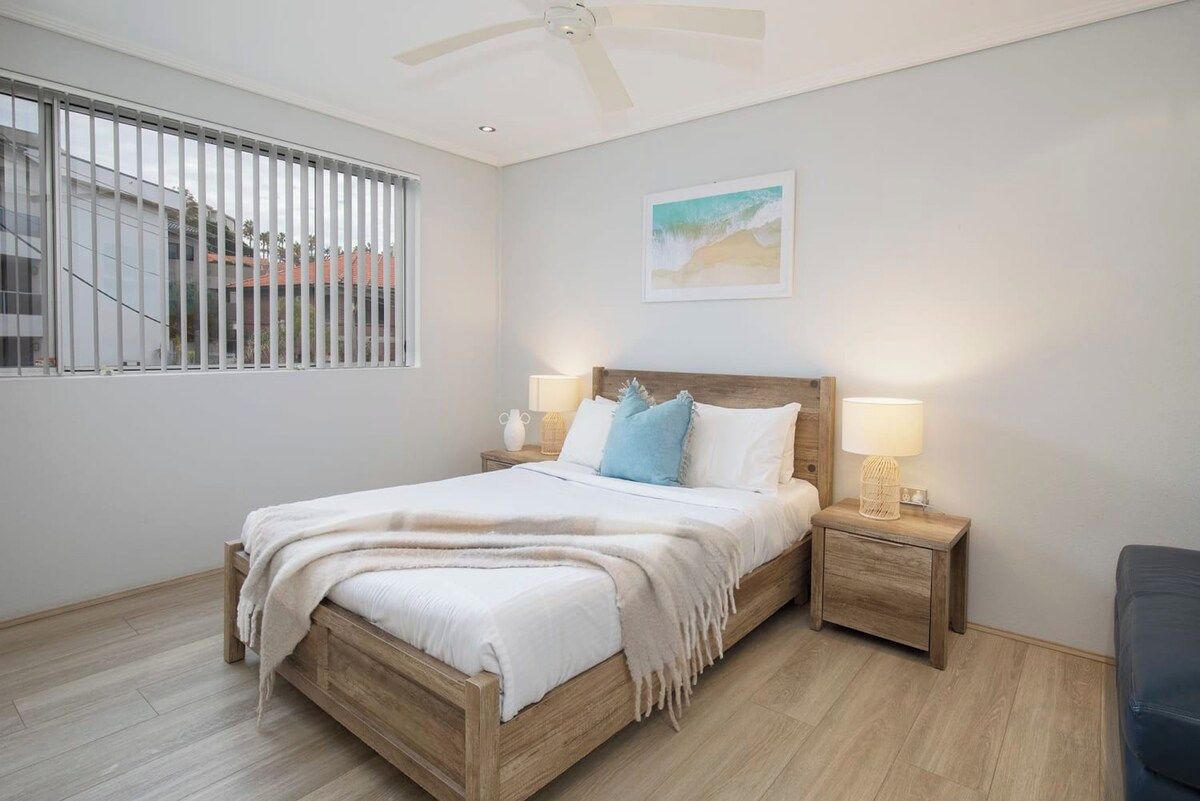 Ein Schlafzimmer mit einem Holzbett, Nachttischlampen, einem Deckenventilator, einem Fenster mit Jalousien und einem gerahmten Bild an der Wand.