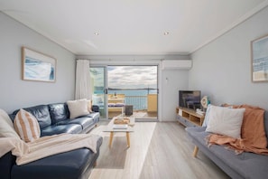 A modern living room with a sofa, a coffee table, and a television. There is a balcony with a view of the sea.