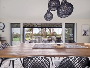 Un espace repas à aire ouverte, avec une grande table en bois, des chaises noires en osier et une vue sur l’extérieur.