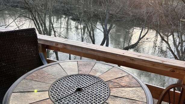 Una mesa redonda con una parrilla metálica en el centro, ubicada en una terraza con barandas de madera que da vista a un río y árboles.