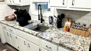 Una cucina con mobili bianchi, un piano di lavoro in granito, un lavello in acciaio inox e diversi utensili ed elettrodomestici da cucina.