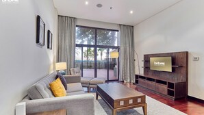 A modern living room with a sofa, coffee table, and TV. There is a balcony with a view of trees and a railing. - 1B-AnantaraSouth-G02 by bnbmehomes (Dubai)