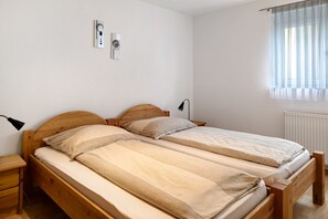 A room with two wooden beds, a nightstand, a lamp, a window with blinds, and a wall-mounted thermostat.
