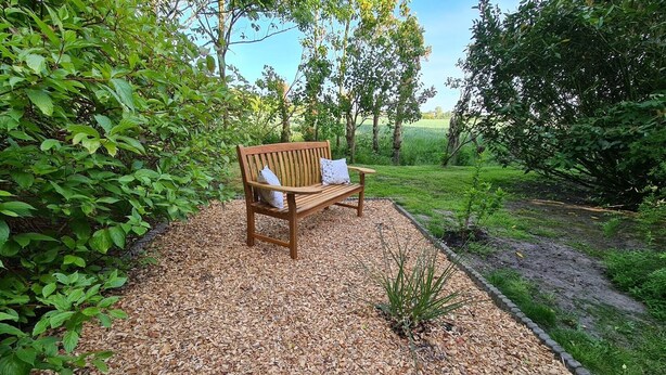 Eine hölzerne Bank mit Kissen in einem Gartenbereich mit Mulch und Steinrand.