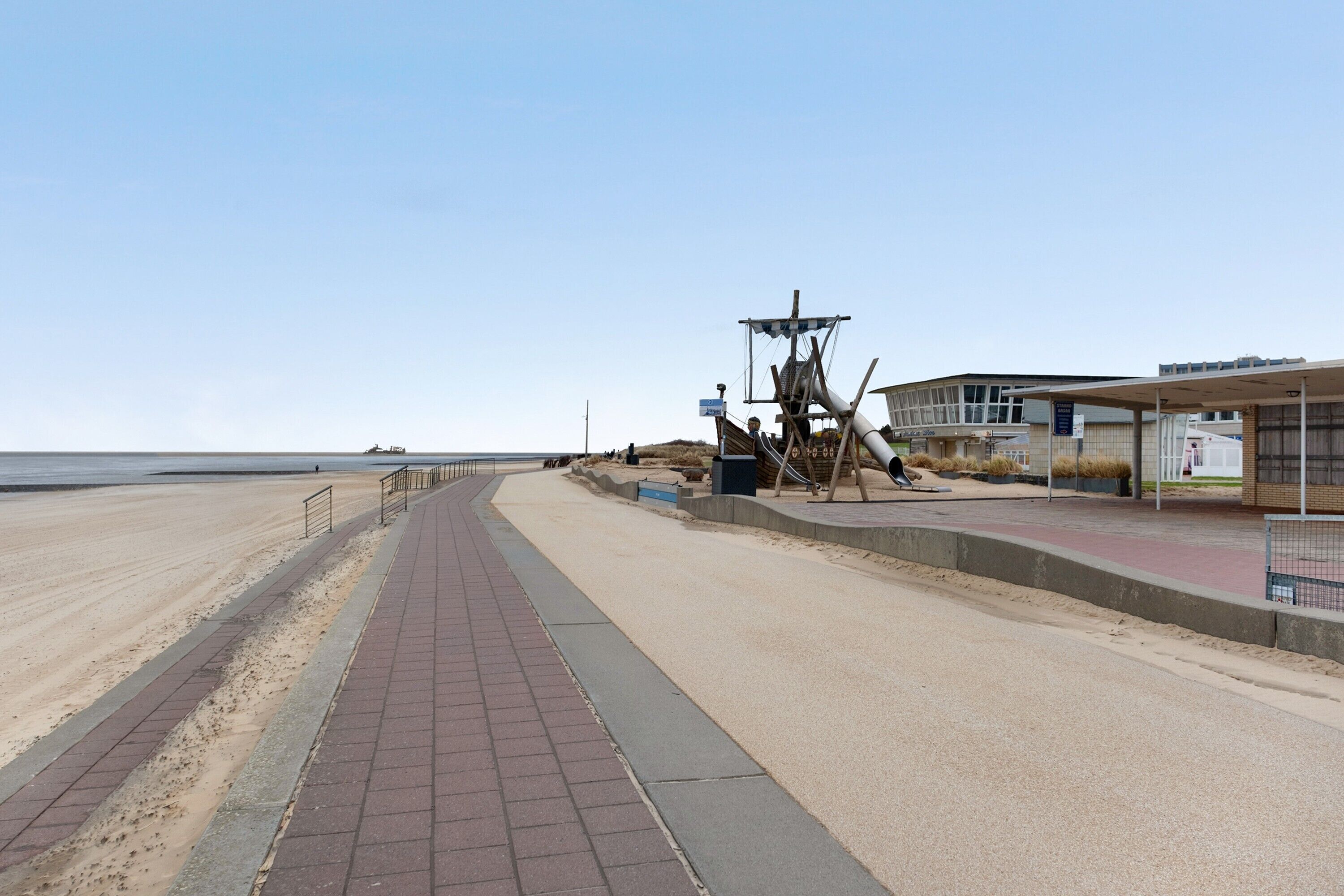 Une promenade en bord de mer avec un sentier de sable, une structure en bois et un bâtiment doté d’une zone abritée.