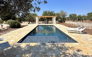 Ein rechteckiger Pool mit Pergola und Liegestühlen auf einer Steinterrasse.