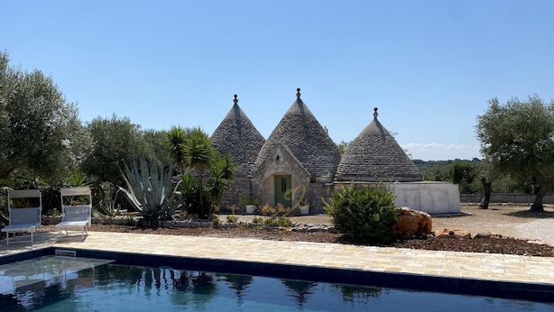 A swimming pool with a stone deck, surrounded by a garden with various plants and a stone building with conical roofs. - TD Trulli Dell Uliveto Cozy Trulli With Pool (Castellana Grotte)