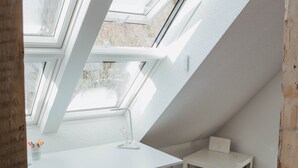 Une pièce avec un plafond en pente, des fenêtres de toit, une chaise noire, une petite table blanche et une poubelle blanche.