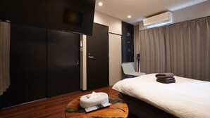 A modern hotel room with a bed, a round wooden table with a towel and remote control, a dark wardrobe, a chair, and an air conditioning unit. - Penthouse 1-bedroom Flat in Hiroo Shibuya (Tokyo)
