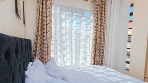 Una cama bien hecha con sábanas blancas en una habitación con un ventanal cubierto por cortinas estampadas.