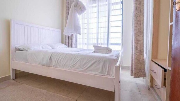 A single bed with white bedding in a room with a window and curtains. - Lux Suites Cavendish Transit  Apartments (Mlolongo)