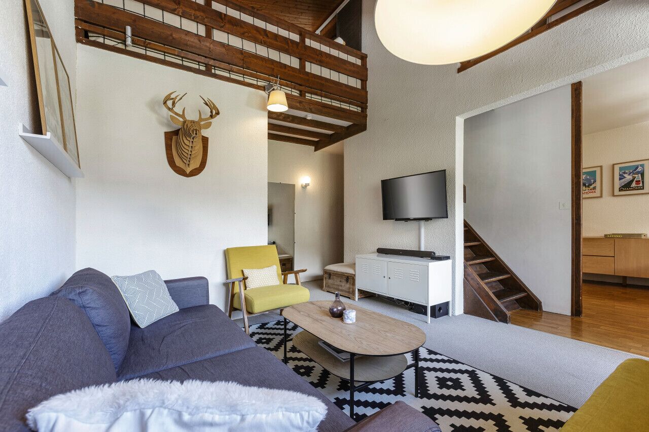 Ein Wohnzimmer mit einer Couch, einem Sessel, einem Couchtisch und einem aufgehängten Hirschgeweih. Ein Fernseher, eine Treppe und eine Holzziel sind ebenfalls vorhanden.