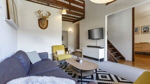 Una sala de estar con un sofá, un sillón, una mesa de centro y una cabeza de venado montada en la pared. Hay un televisor, una escalera y un techo de madera.