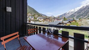 Un balcón de madera con una mesa y sillas, con vista a un pueblo de montaña con casas tradicionales y picos nevados.