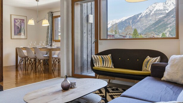 Una sala de estar moderna con sofá, mesa de comedor y sillas. Un amplio ventanal ofrece vistas a la montaña.