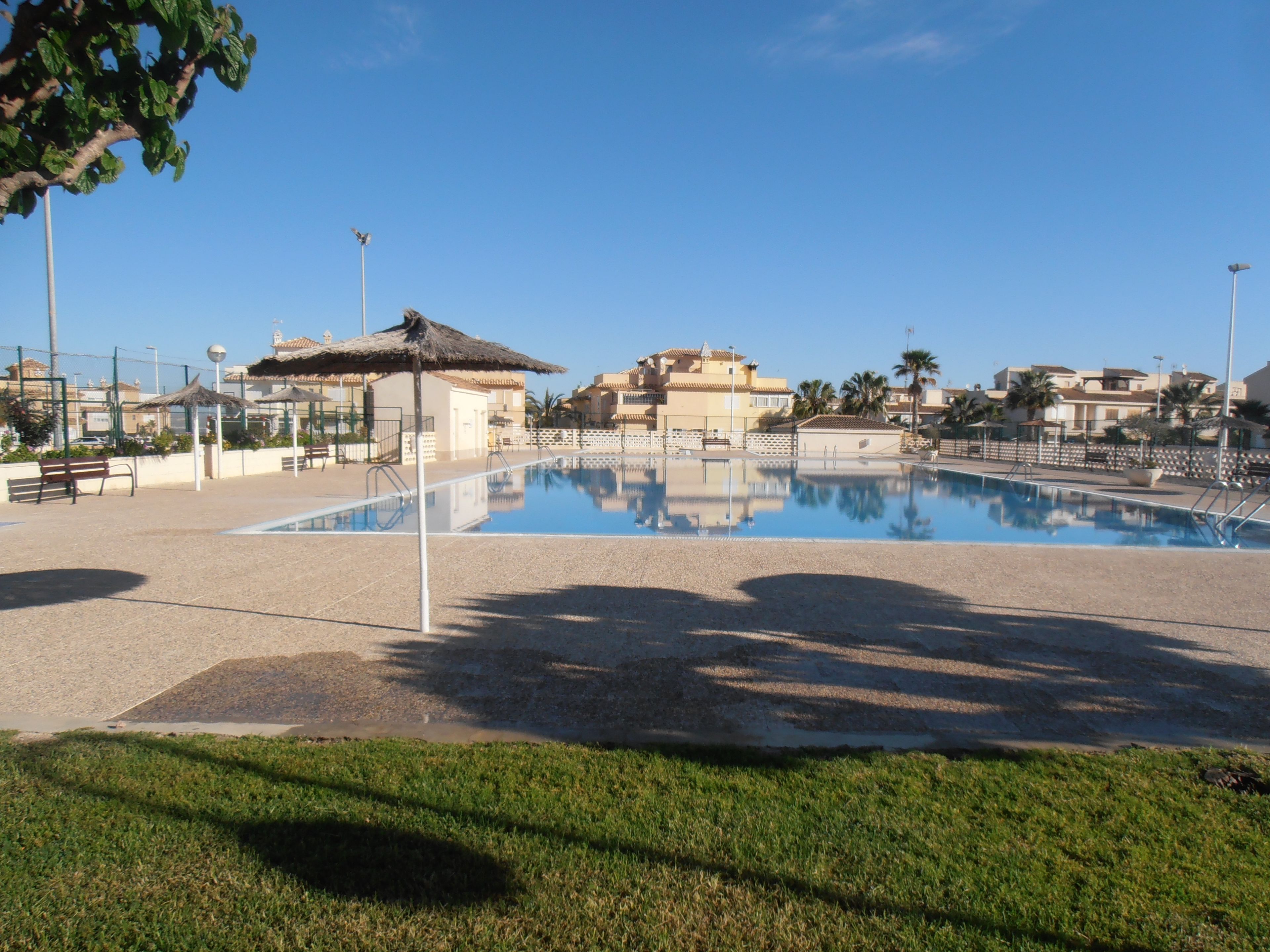 A swimming pool with a clear blue sky, surrounded by a paved area with benches and a small building with a thatched roof.