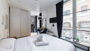 A hotel room with a bed, a desk, a chair, a small table with a plant, and a view of the city through large windows.