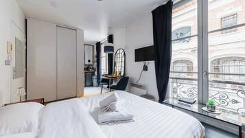 Une chambre d’hôtel avec un lit, un bureau, une chaise, une petite table avec une plante et une vue sur la ville grâce à de grandes fenêtres.