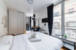 A hotel room with a bed, a desk, a chair, a small table with a plant, and a view of the city through large windows.