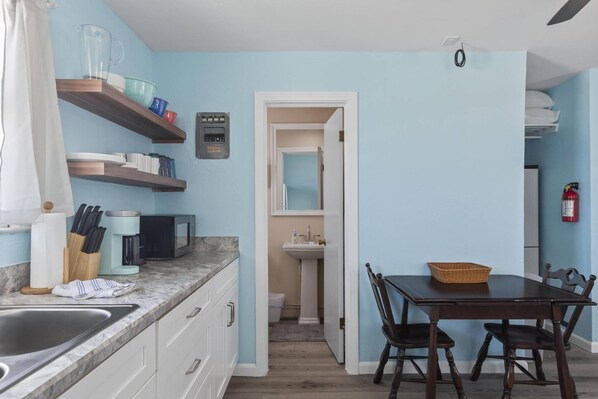 Una cocina con encimera de mármol, un lavamanos y una zona de comedor con mesa y sillas. Hay una estantería con vajilla y un microondas en la encimera.