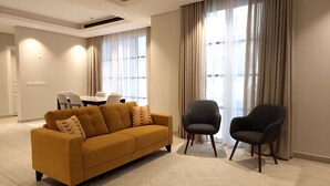 Una sala de estar moderna con un sofá y dos sillones, una zona de comedor con una mesa y sillas, y ventanas amplias con cortinas.