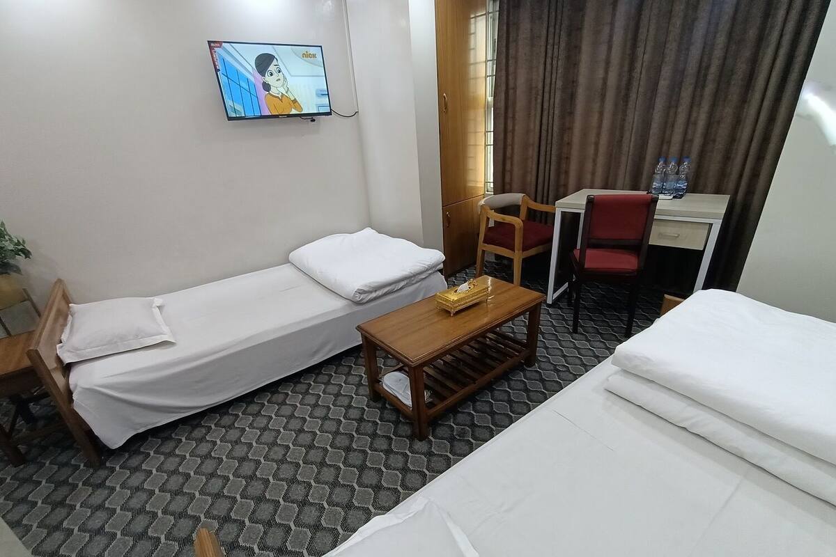 A hotel room with two single beds, a wooden table, a TV mounted on the wall, a desk with a chair, and a window with curtains.