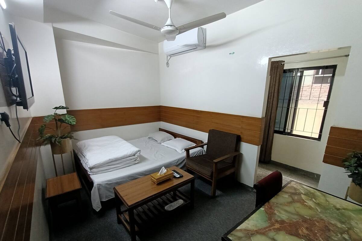 A compact hotel room with a single bed, a wooden table, a chair, a small desk, and a ceiling fan.