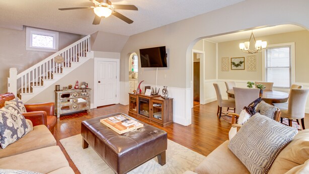 A spacious living room with a fireplace, a dining area with a chandelier, and a staircase with a ceiling fan.