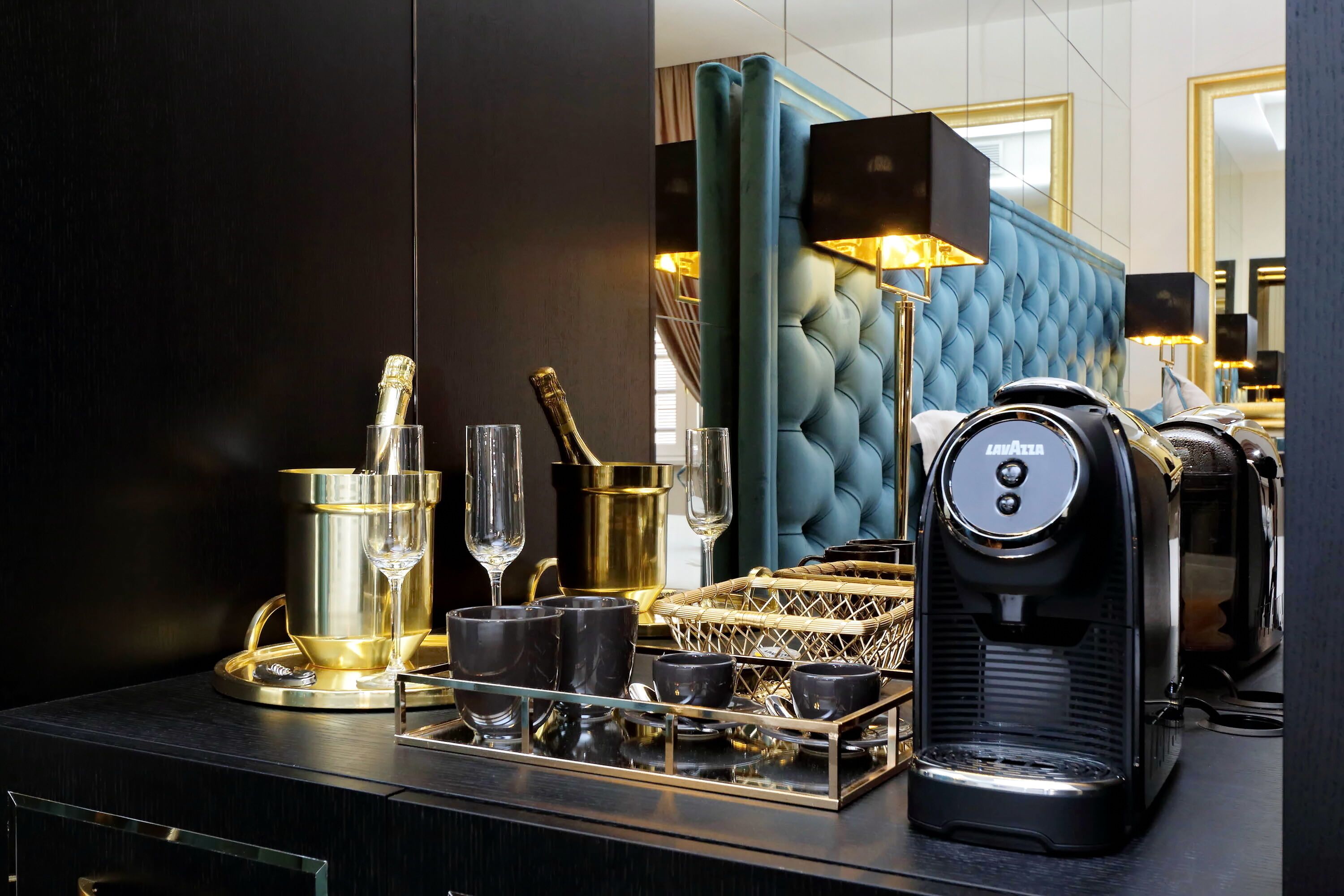 A luxurious hotel room with a dark wood finish, a tufted blue headboard, a coffee maker, and a tray with champagne glasses and cups.