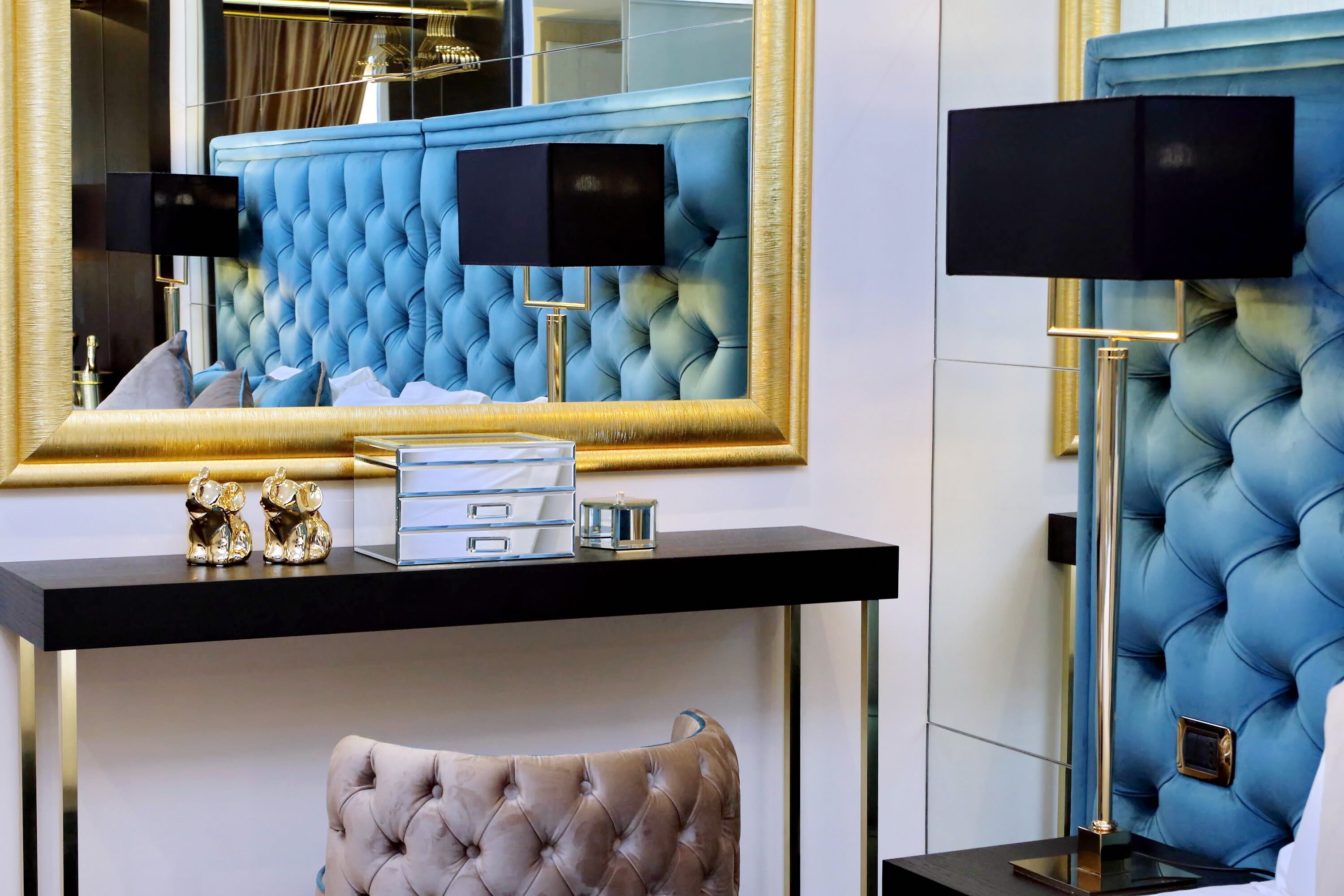 A luxurious room with a tufted blue headboard, a large mirror, a black console table with drawers, a tufted armchair, and a floor lamp.