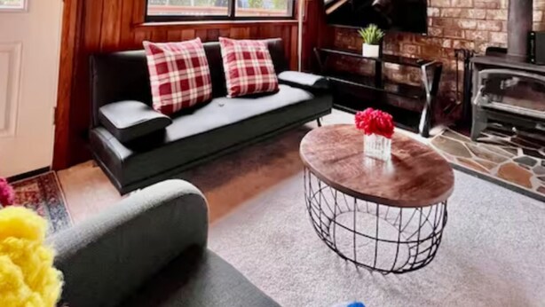 A cozy living room with a wooden ceiling, a fireplace, a sofa with plaid cushions, a round coffee table, and a large window offering a view of greenery. - Unique/modern/Mt Si/Shopping (North Bend)