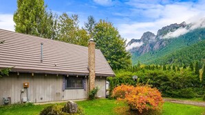 A wooden cabin with a chimney, surrounded by lush greenery and a mountain backdrop. - Unique/modern/Mt Si/Shopping (North Bend)