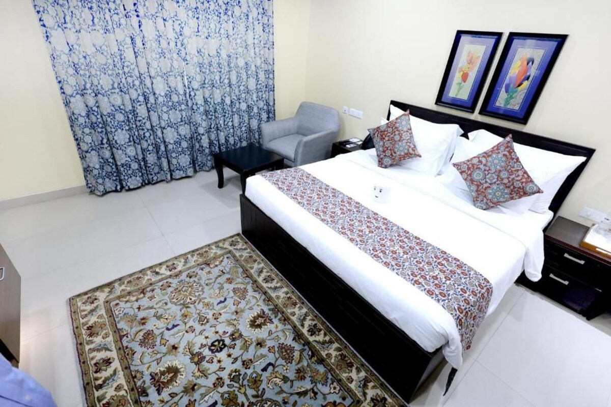 A bedroom with a bed, a chair, a nightstand, a desk, a patterned rug, and a curtain.