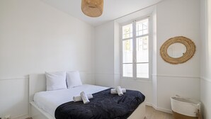 A clean, white bedroom with a bed, a window, a woven wall decoration, and a wicker basket. - Le Windsor avec extérieur, pour un plaisir partagé (Cannes)