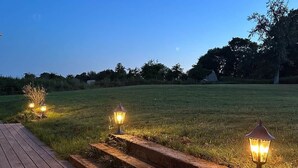 Eine Holzterrasse mit Stufen, die zu einer Rasenfläche führen, beleuchtet von drei Laternen.