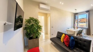 Una sala de estar moderna con un sofá negro, una planta grande en maceta y una zona de comedor con una mesa dispuesta para dos personas.