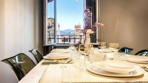 Una mesa de comedor dispuesta con platos, vasos y cubiertos, con vista a un paisaje urbano a través de una ventana abierta.