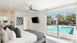 Un dormitori modern amb un llit gran, un ventilador de sostre, una televisió de pantalla plana i vista a una piscina a través de grans portes corredisses.