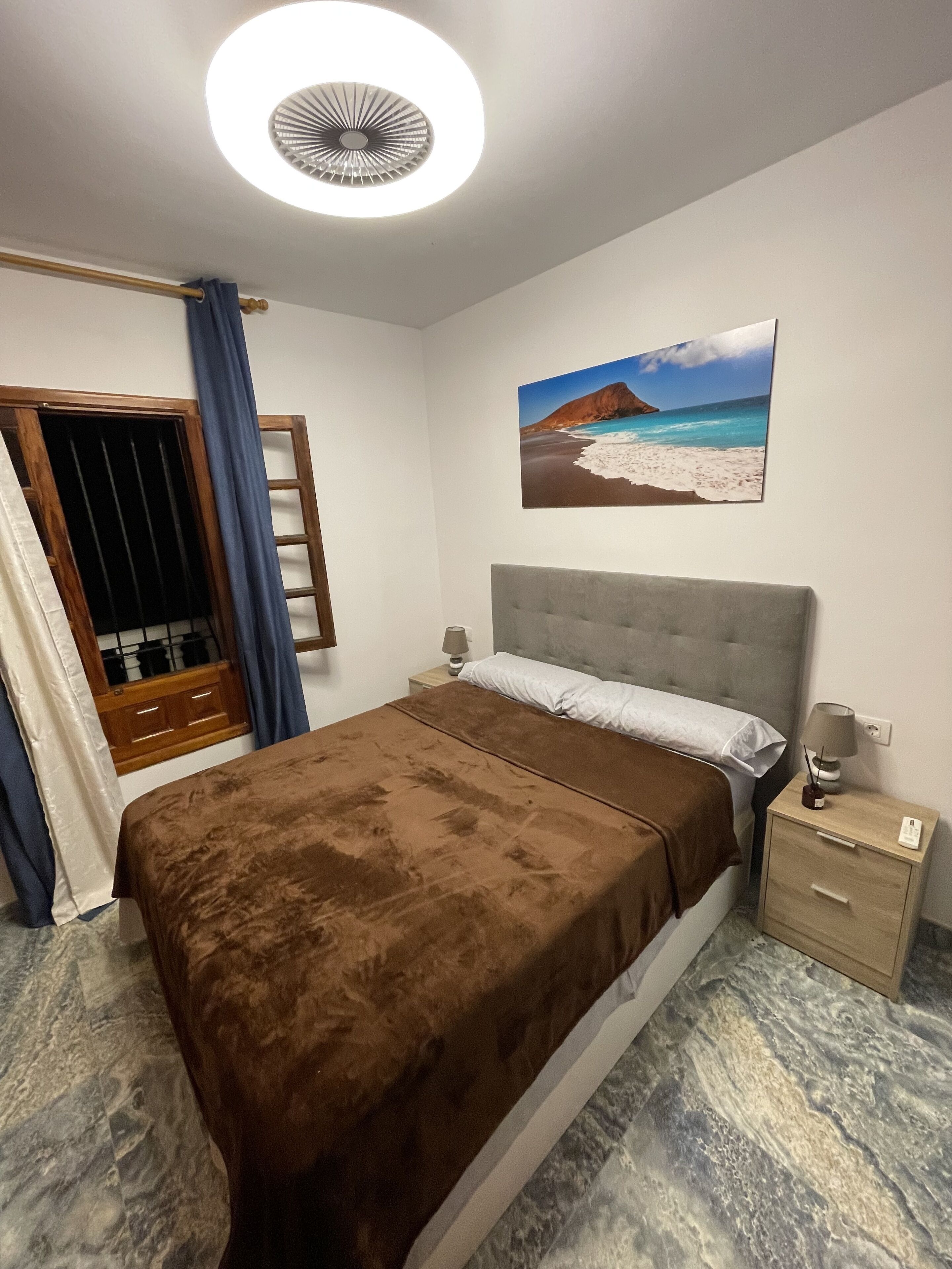 A bedroom with a bed, a nightstand, a wall-mounted lamp, a ceiling fan, a window with curtains, and a beach scene painting.