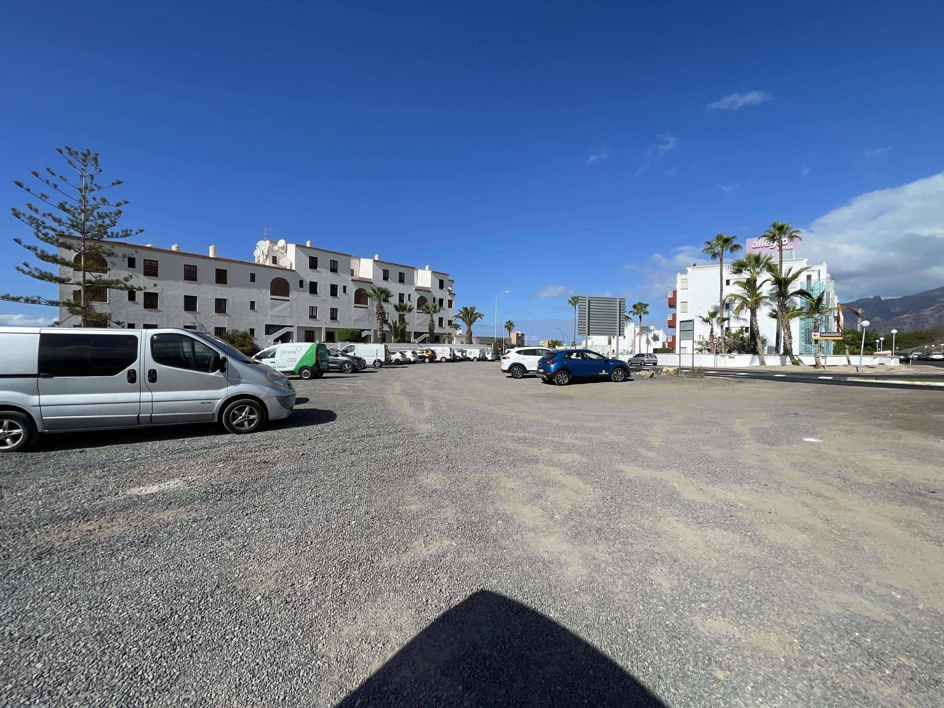 A parking lot with several vehicles, including a silver van and a blue car. There are palm trees and a building with a sign that reads "Varadero" in the background.
