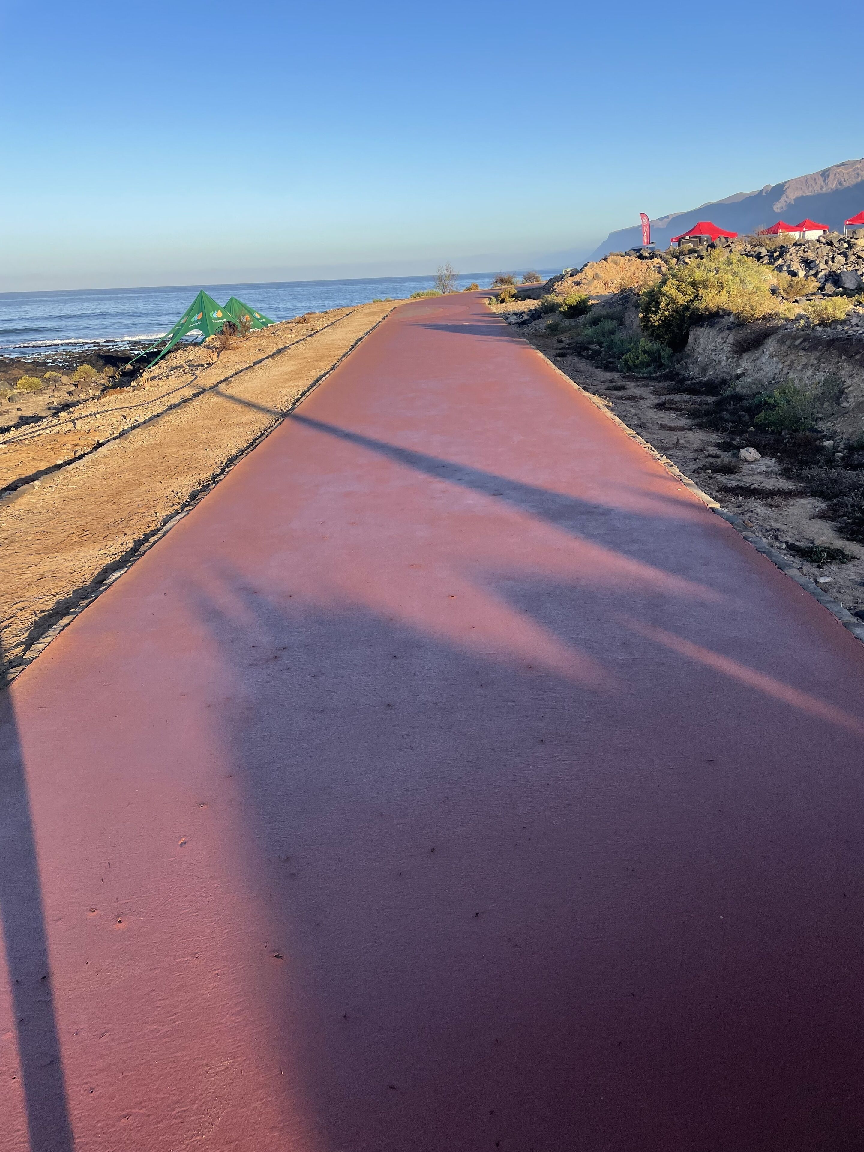 A paved pathway along a coastal area with red tents and a flag on the right, leading to the sea.