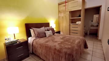 Un dormitorio con cama, mesitas de noche, lámpara, armario y baño al fondo.