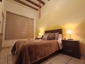 Ein Schlafzimmer mit einem Bett, zwei Nachttischen, einer Lampe auf jedem Tisch, einem Fenster mit Jalousien und einem Holzbalken an der Decke.