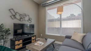 A modern living room with a flat-screen TV, a sofa, a coffee table, and a large window with blinds. - Des Moines Designer 1BD 1BA Downtown Apartment (Des Moines)