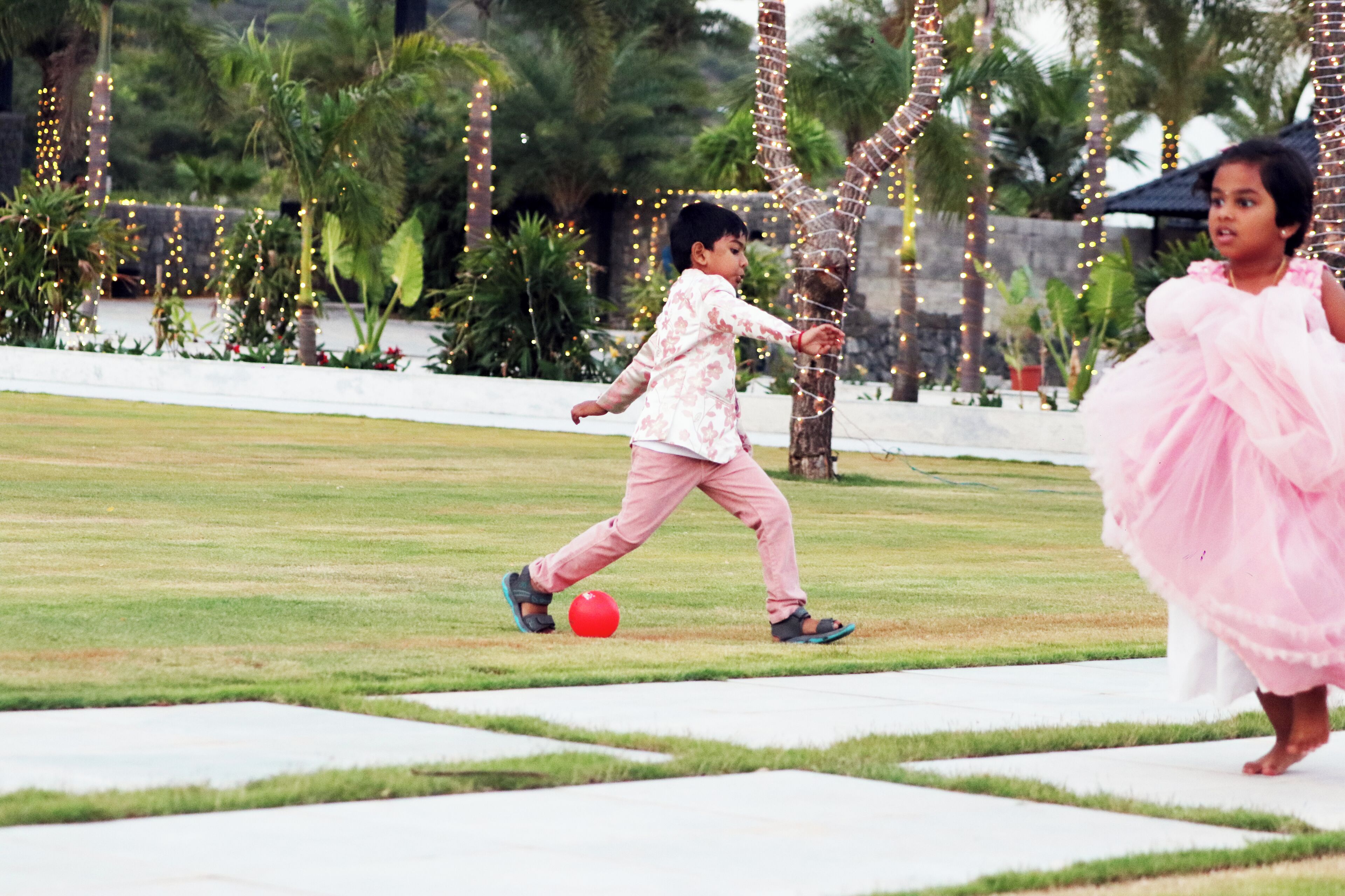 A child playing with a red ball in a grassy area with decorative lights and palm trees.