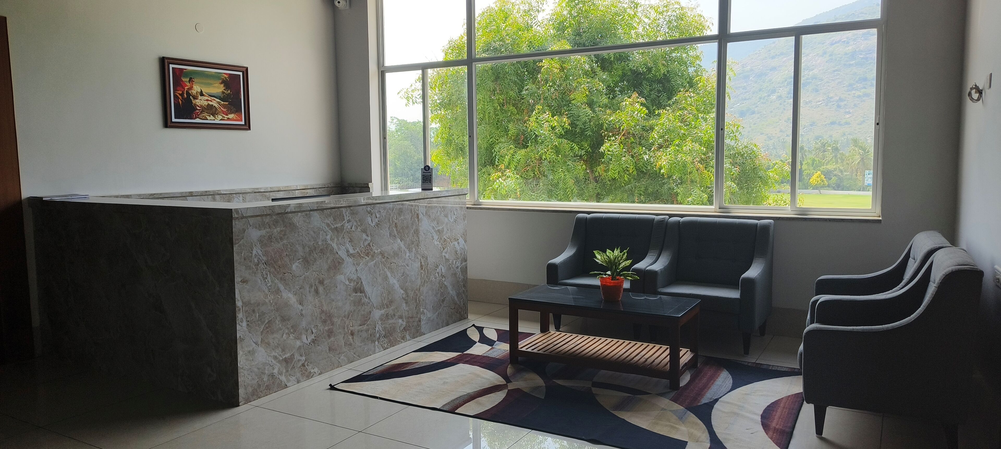 A reception area with a marble counter, a framed painting, a seating area with a sofa and chairs, a coffee table with a plant, and a view of greenery through large windows.