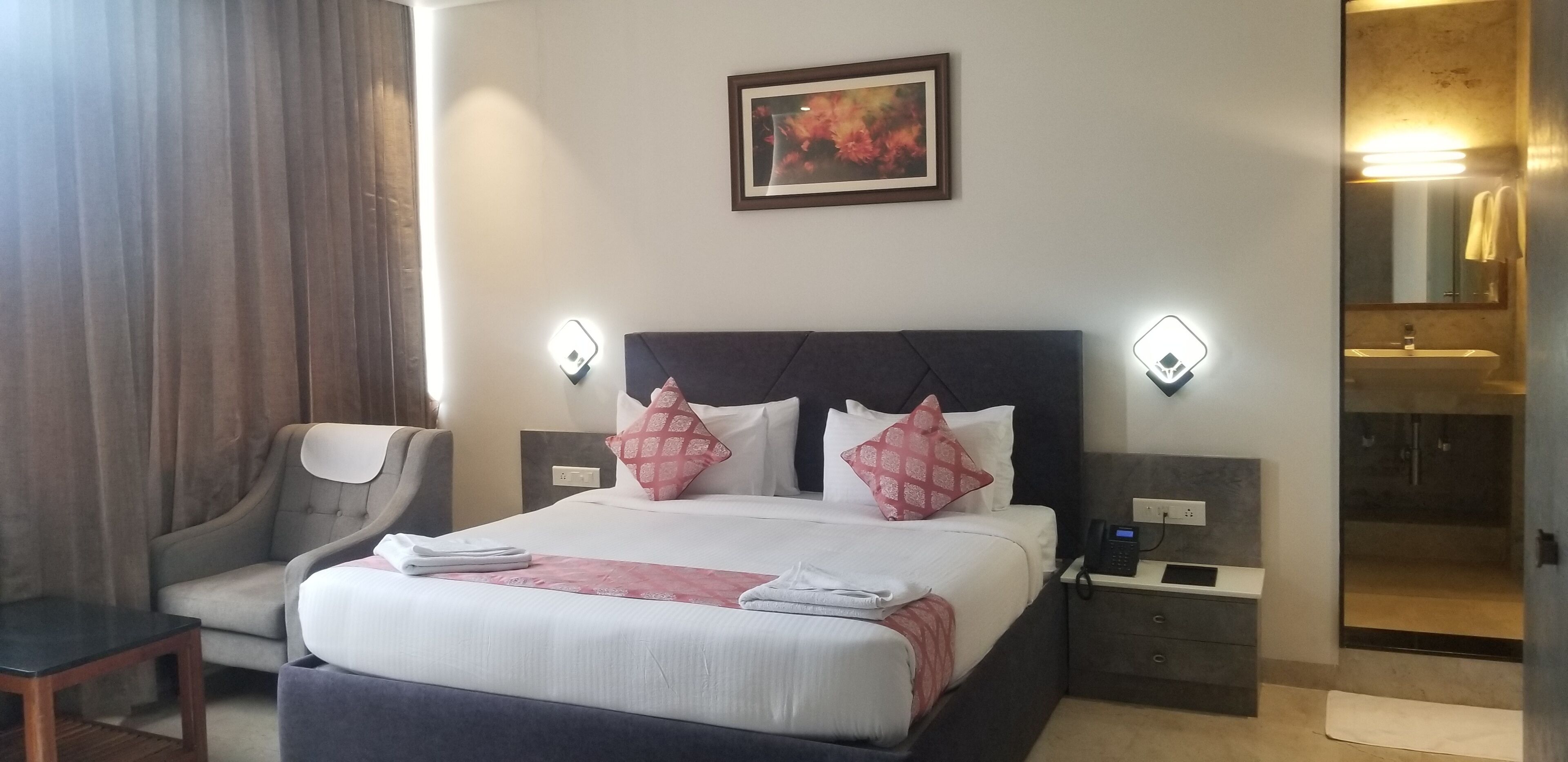 Habitación de hotel con cama, una silla, mesita de noche con teléfono y baño visible a través de una puerta abierta.