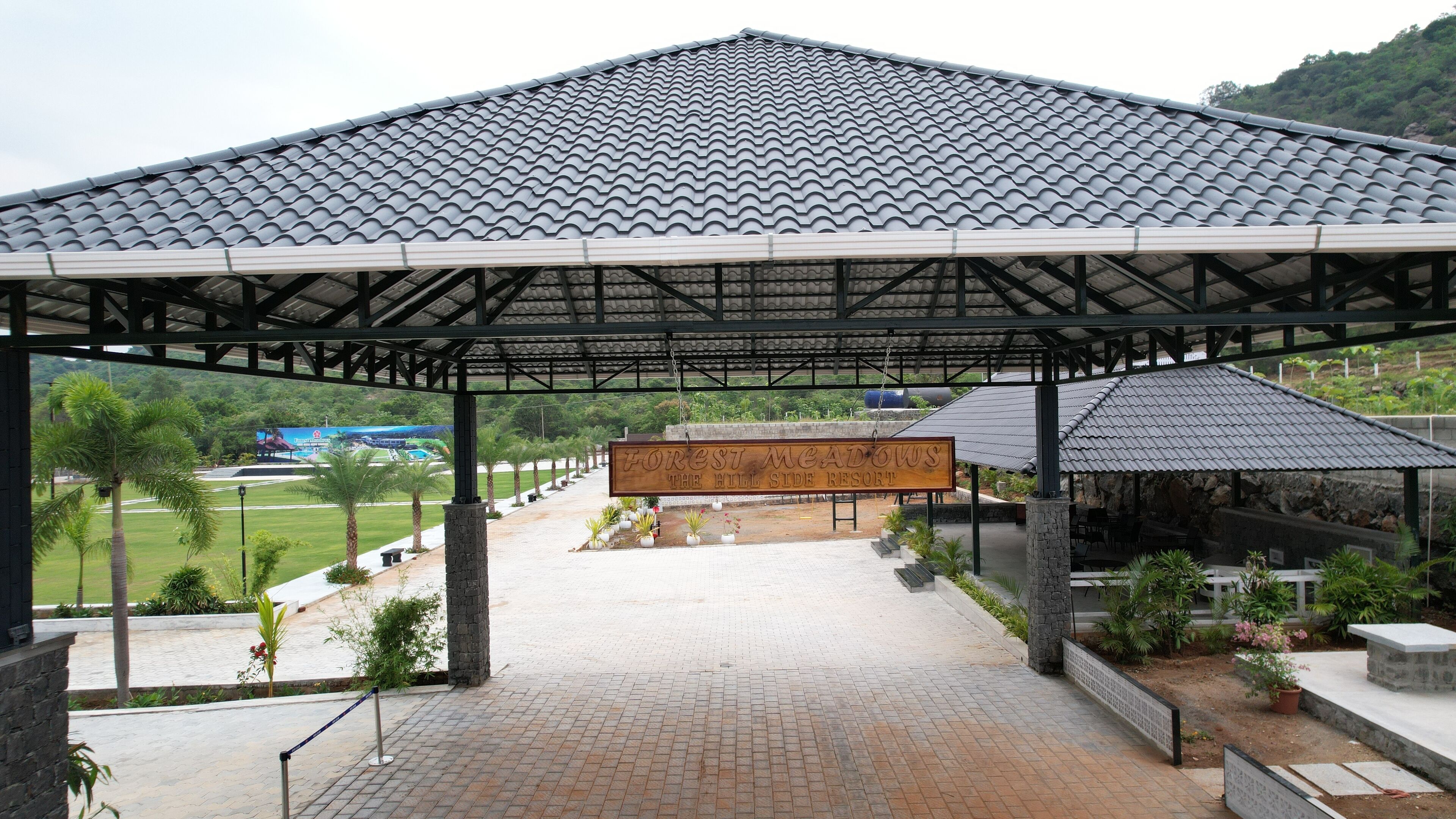 Un pabellón cubierto con techo de tejas, un cartel que dice "Forest Meadows The Hill Side Resort" y un camino pavimentado que lleva a una zona verde con palmeras.