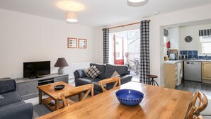 Una sala de estar moderna con sofá, televisor y mesa de comedor. Se ve una cocina con pileta y electrodomésticos a través de una puerta abierta.